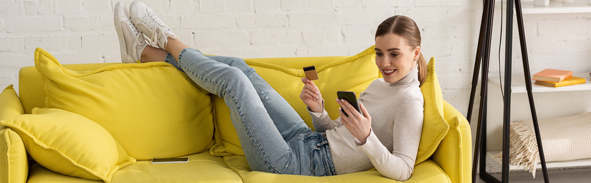 A young woman is lying on her couch enjoying shopping with her phone and credit card.