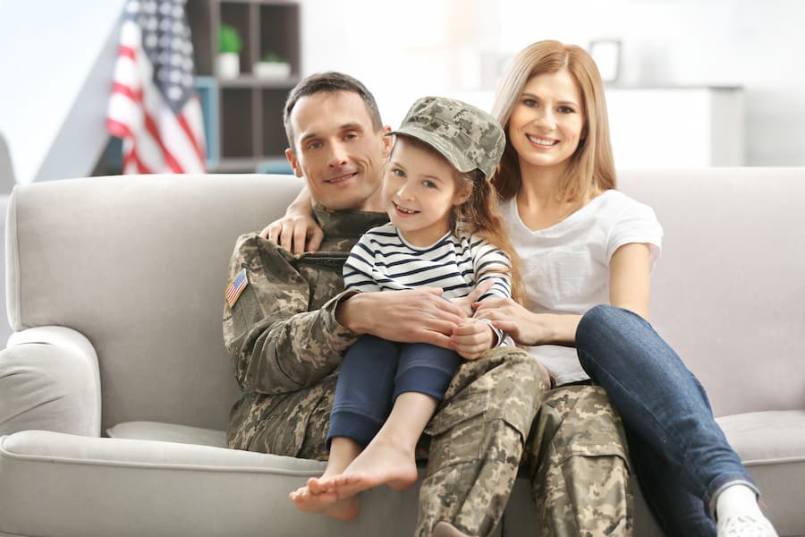 A soldier in uniform sits with his wife and young daughter on a sofa at home