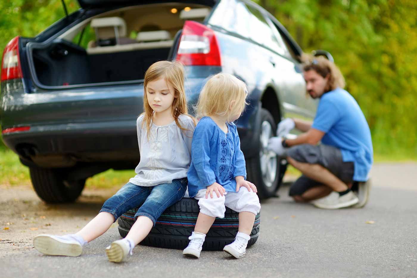Father works on car on side of the road while two daughters sit sadly on tire