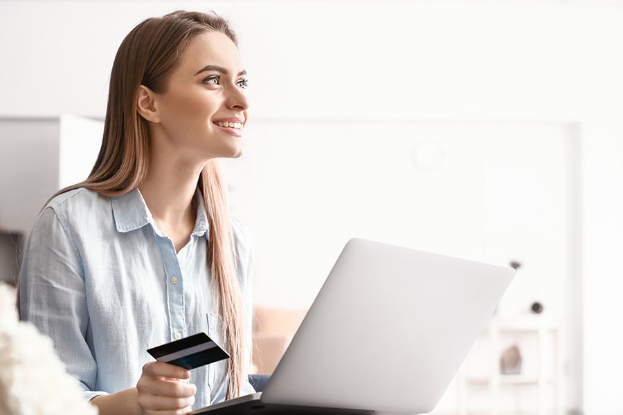 A young woman is holding her debit card and looking at her checking account balance on her laptop.