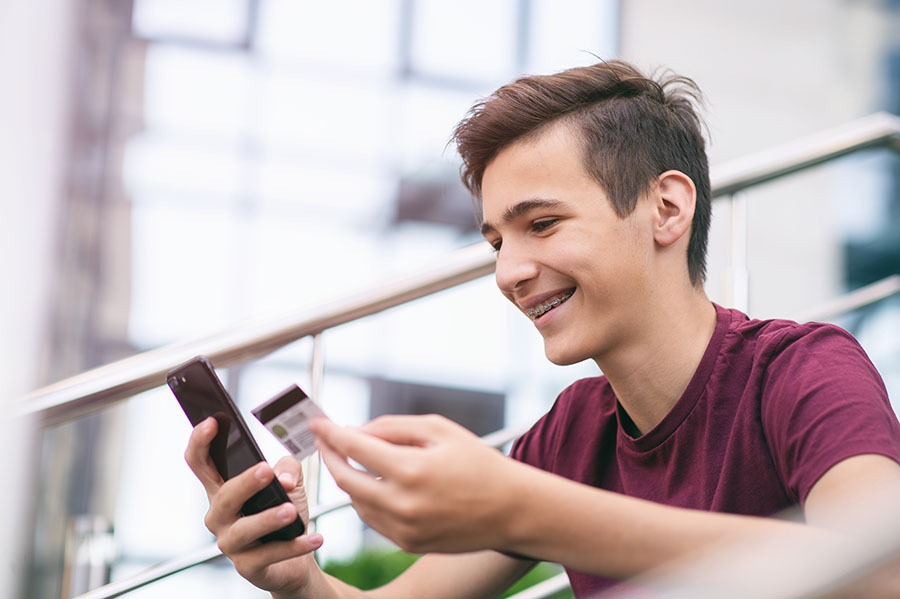 A teenaged boy is holding his debt card and looking at his checking account balance on his phone.