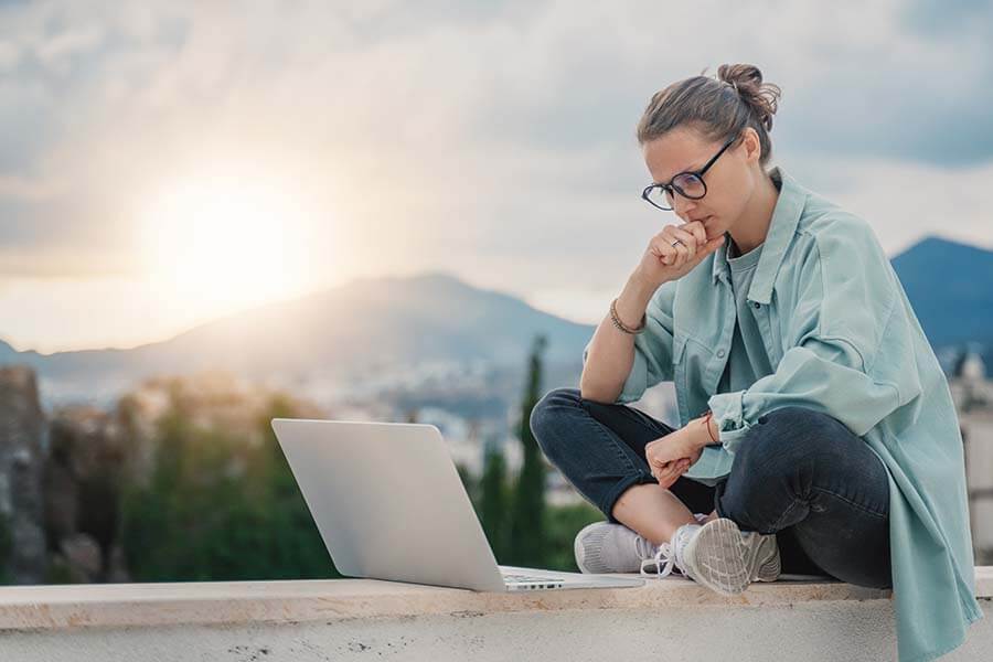 A young woman sitting outside doing research on her laptop to see if she can add money to a certificate of deposit regularly.