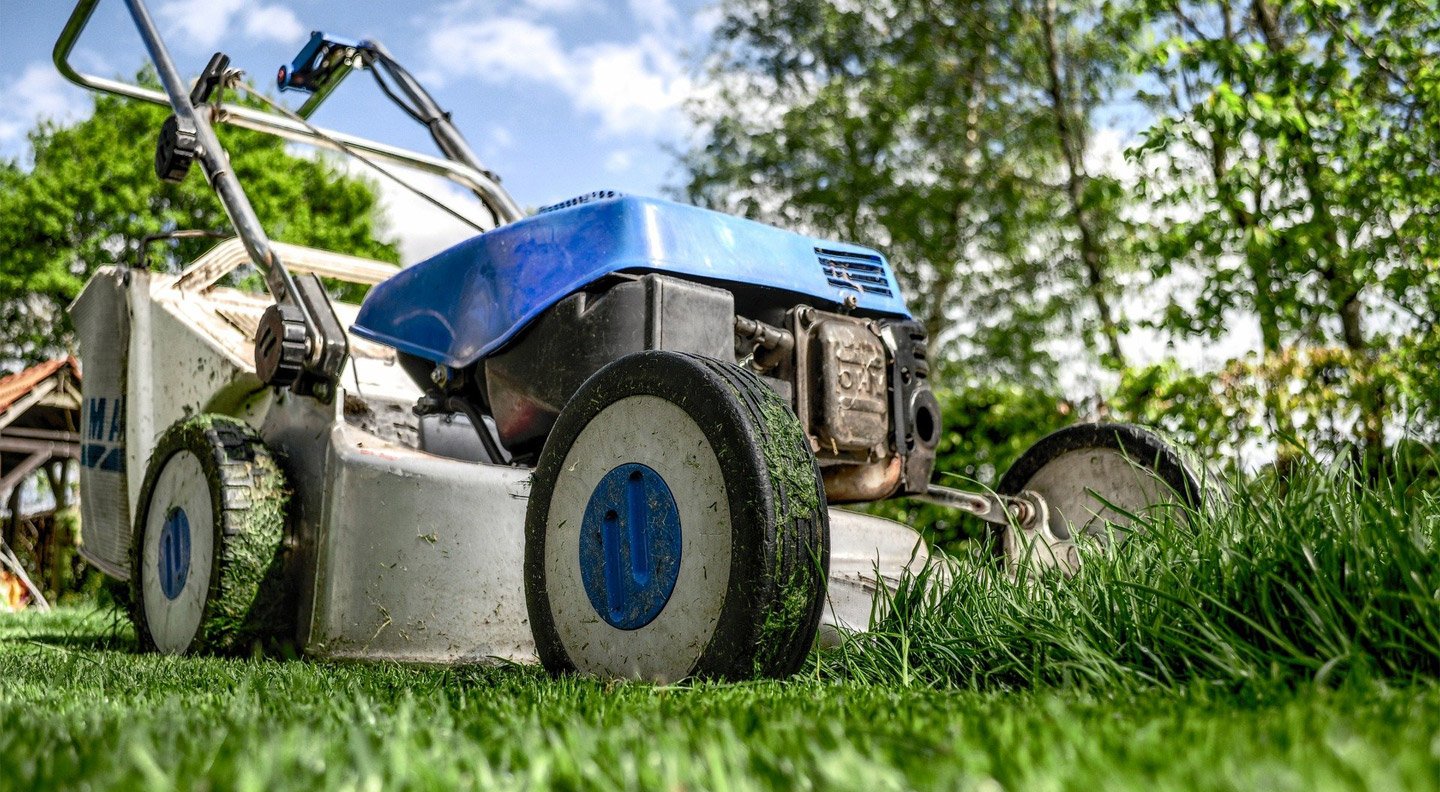 a blue, gas push lawnmower cutting green grass in a lawn with trees and a blue sky in the background