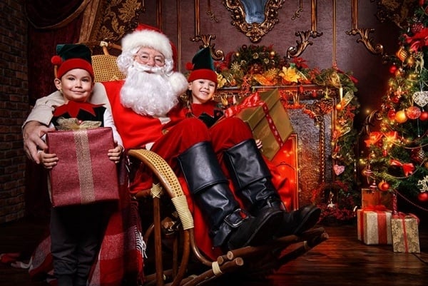 Santa sitting with an elf holding a present on either side of him.