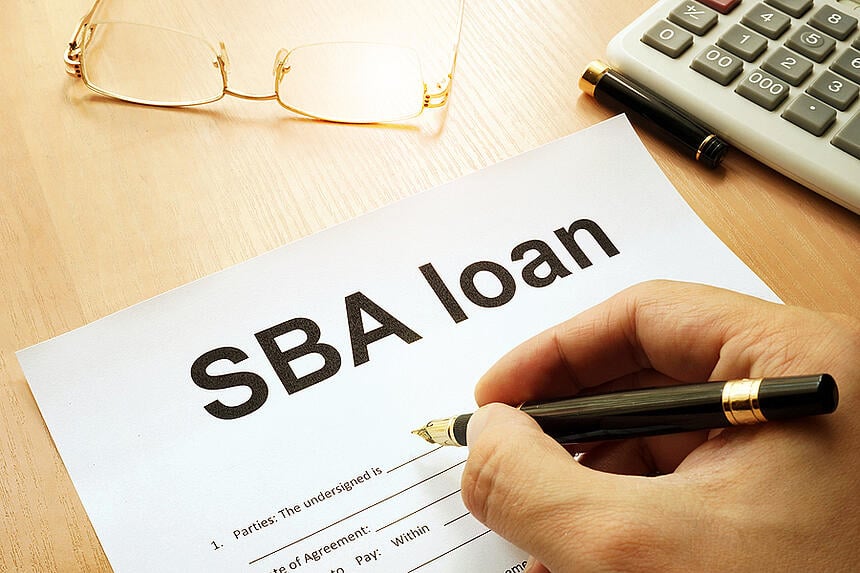 a close up view of a person's hand filling out a paper SBA loan application on a desk with eyeglasses and a calculator nearby