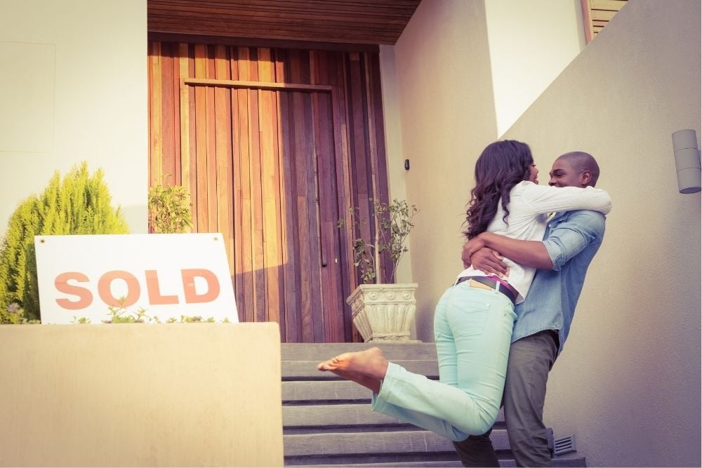 A happy African American couple hugs in front of their new house as first-time home buyers